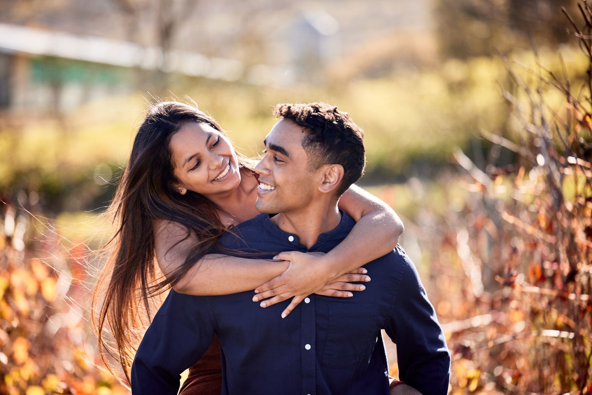 Couple embracing outdoors in warm sunlight, smiling amid autumn foliage.