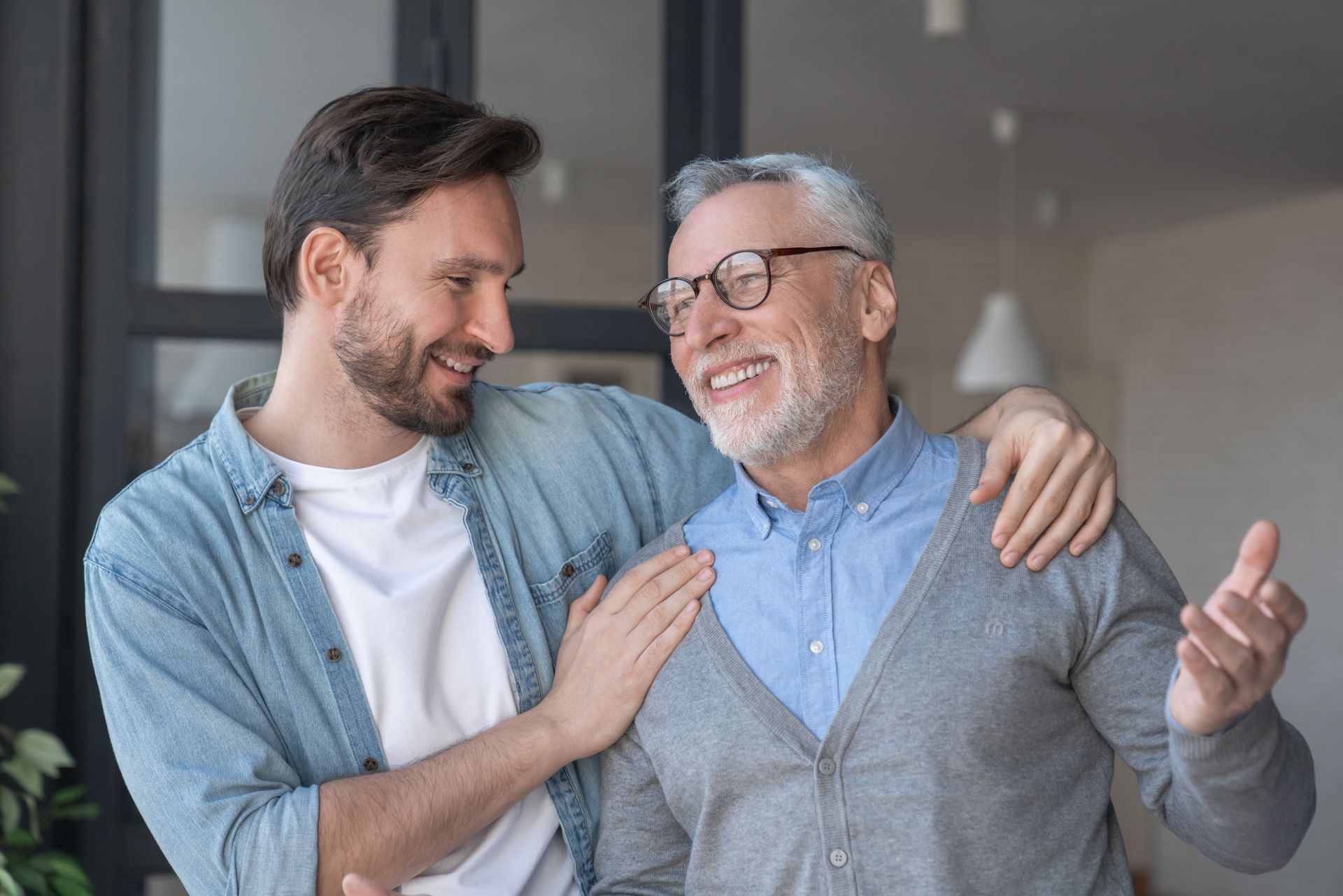 Two smiling men stand together indoors, one with an arm around the other’s shoulder.