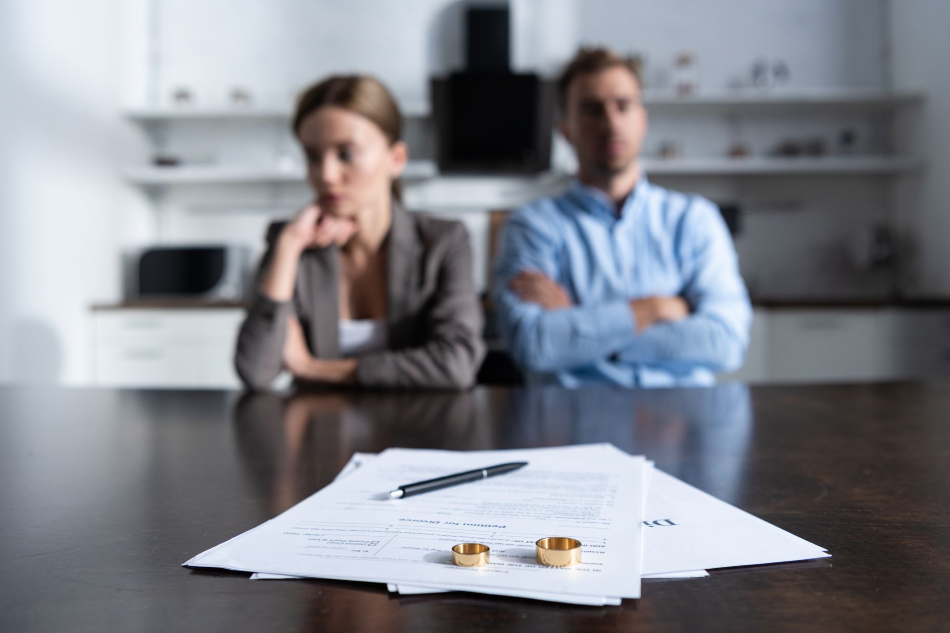 Unsigned divorce papers with wedding rings on a table, with a blurred couple sitting apart in the background
