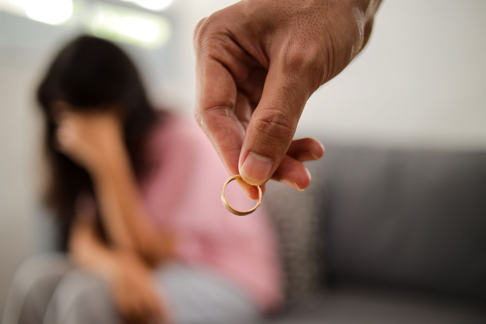 Hand holding a gold ring in front of a blurred woman sitting on a couch