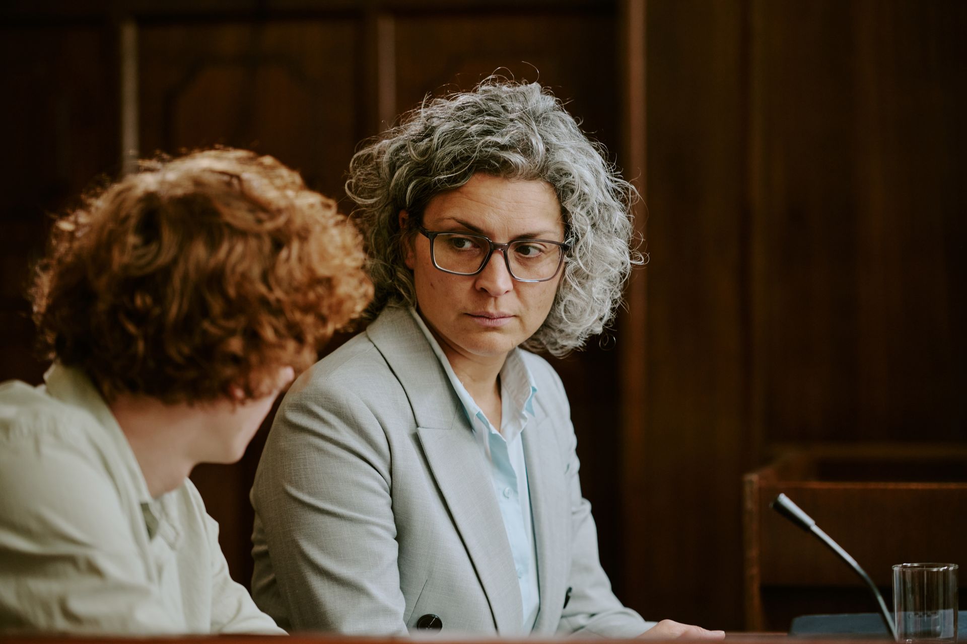 Two people seated at a table in a wood-paneled meeting room, one looking toward the other.