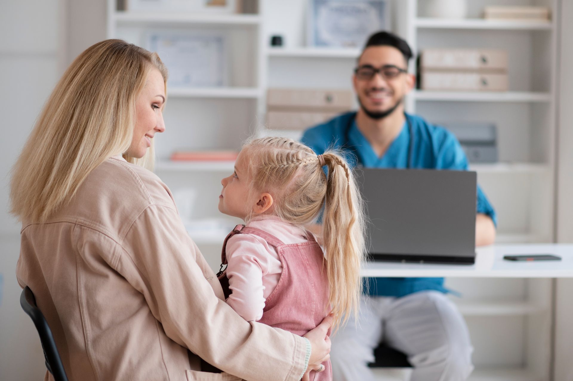 Pediatric care professional sitting behind a desk at his computer with a mother holding her daughter facing him in an office.