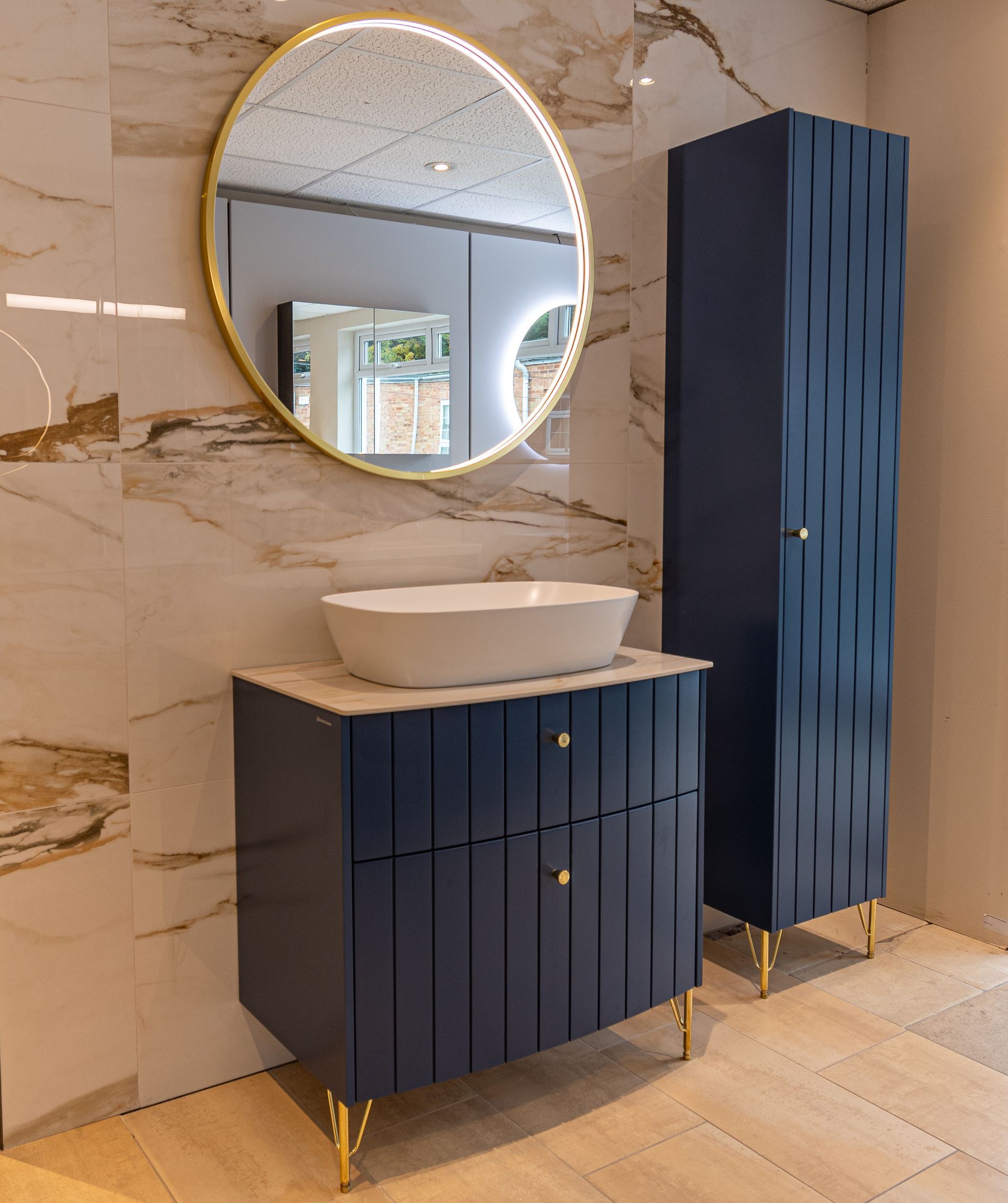 Alternate angle of a bathroom display with a round mirror framed in gold, rectangular countertop sink, and blue paneled-style counter with storage