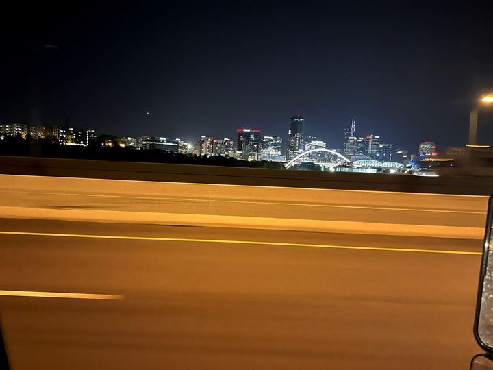 City skyline at night seen from a moving road, with blurred orange barriers in the foreground.