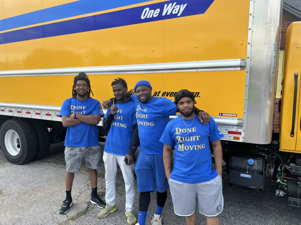 Four people in blue shirts stand beside a yellow moving truck labeled “One Way.”