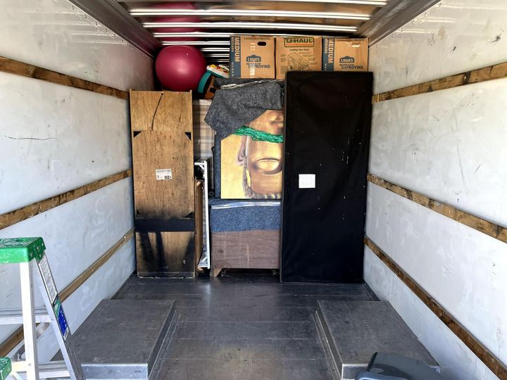 Inside a moving truck with packed furniture, boxes, and a ladder near the open rear door