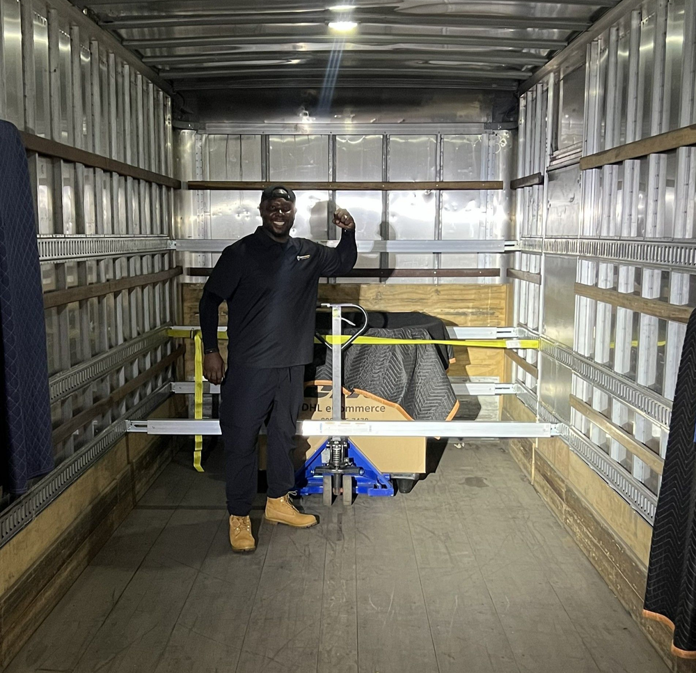 Man standing in a metal shipping container beside a wheeled cart and yellow safety railings
