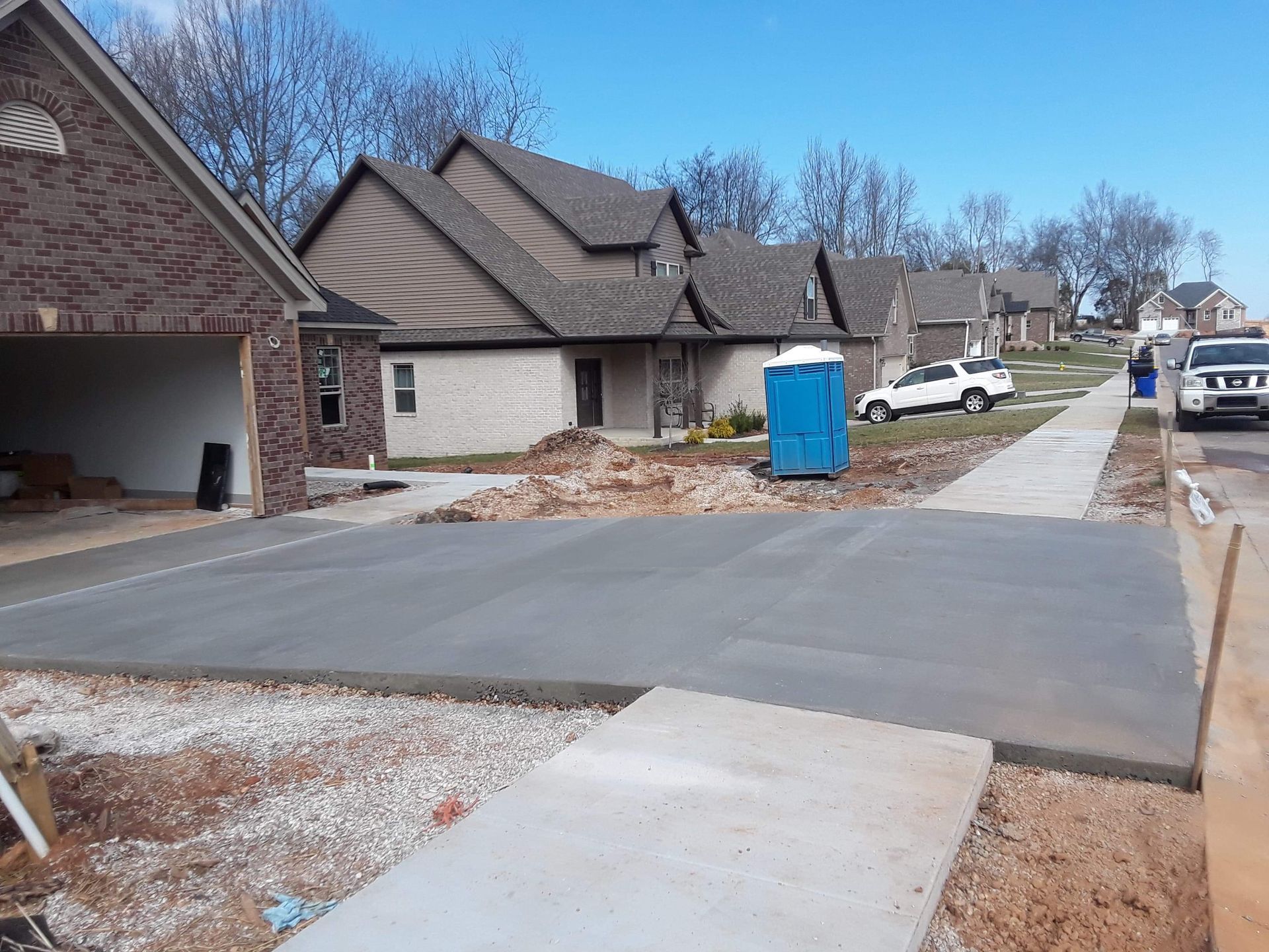 New suburban driveway and sidewalk beside beige houses, with a blue portable toilet and parked truck nearby