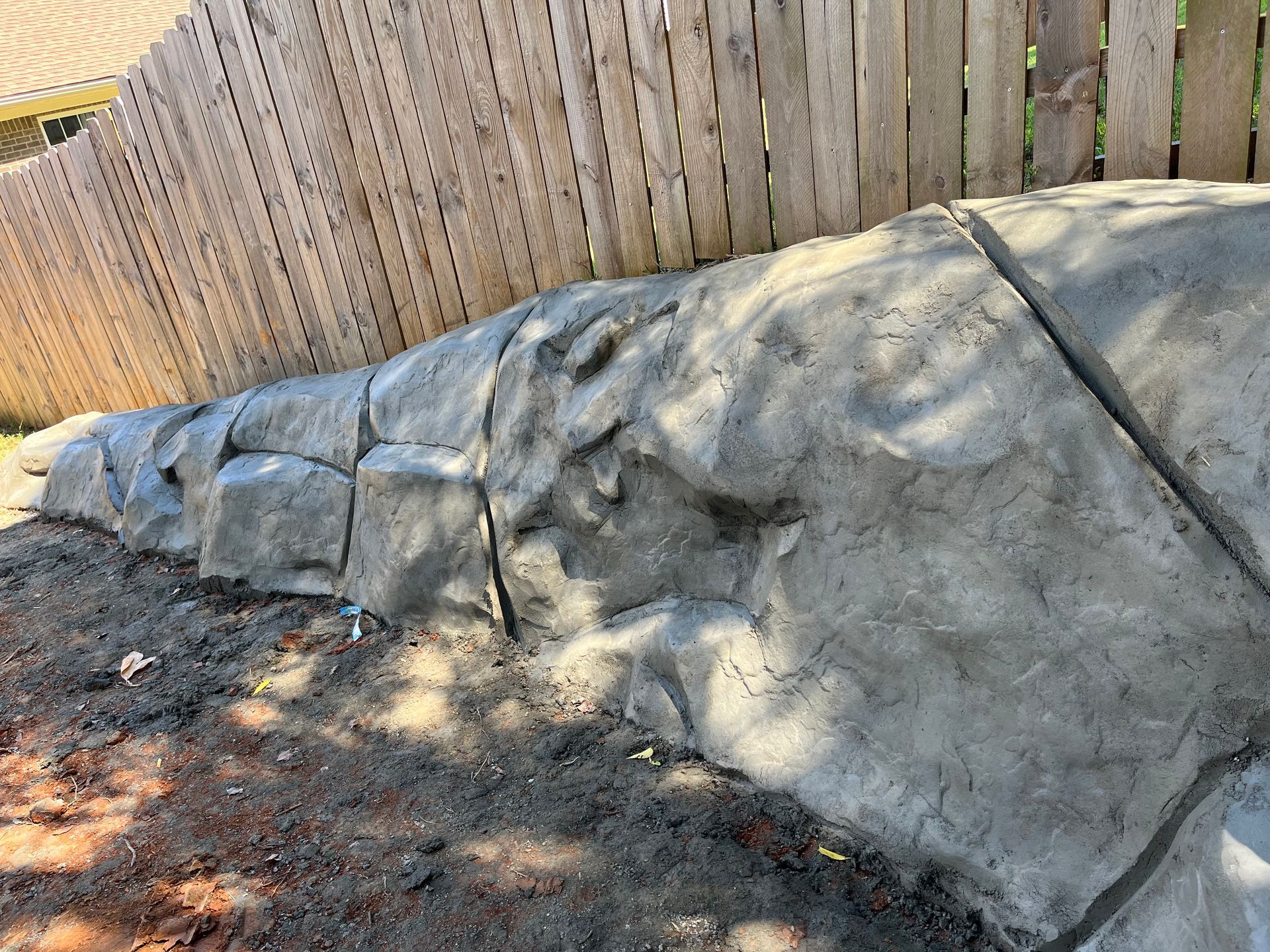 Large gray rock retaining wall beside a wooden fence in a dirt yard, with shadows from trees.