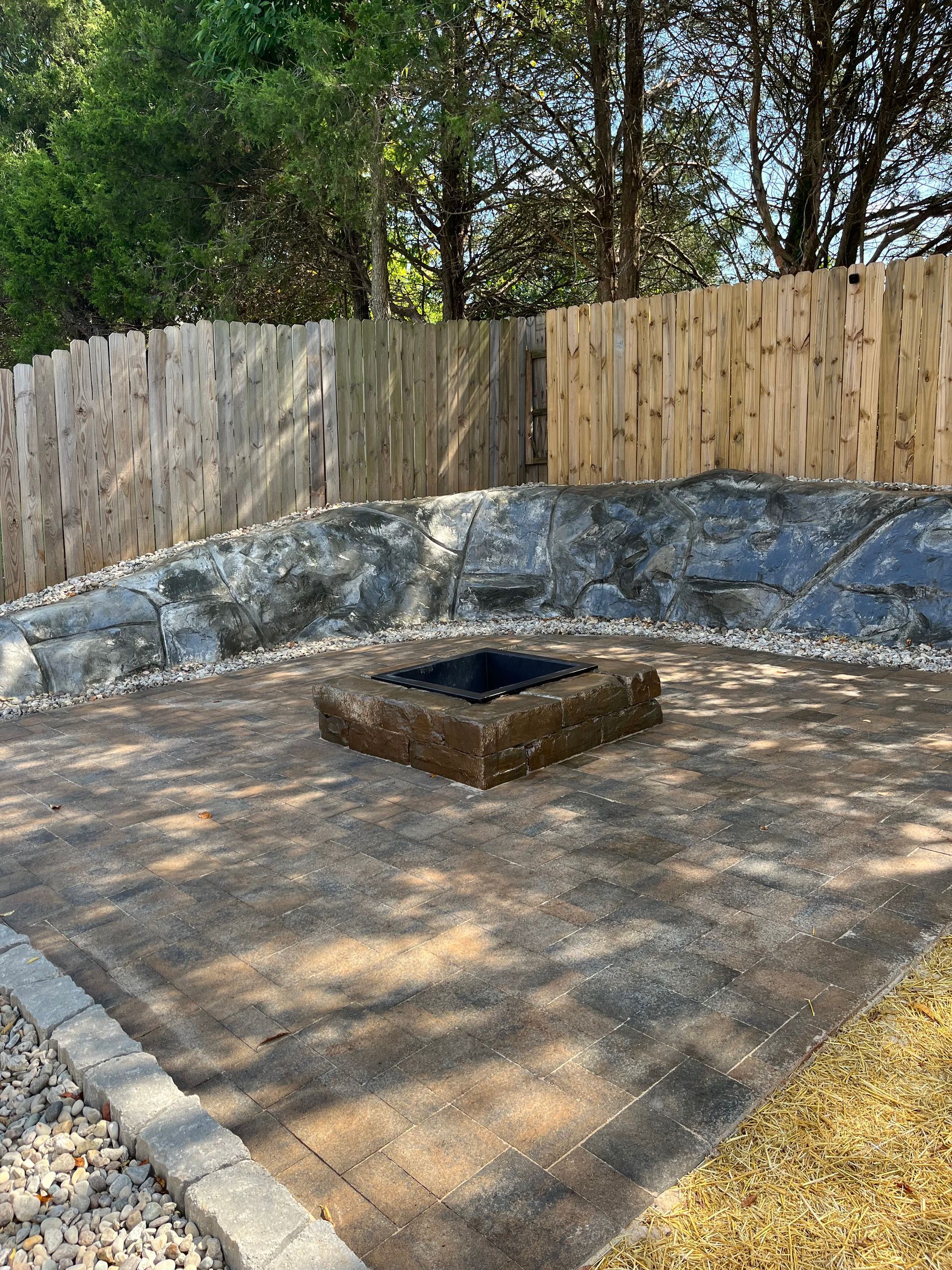 Outdoor stone patio with a square fire pit, wooden fence, and trees in the background