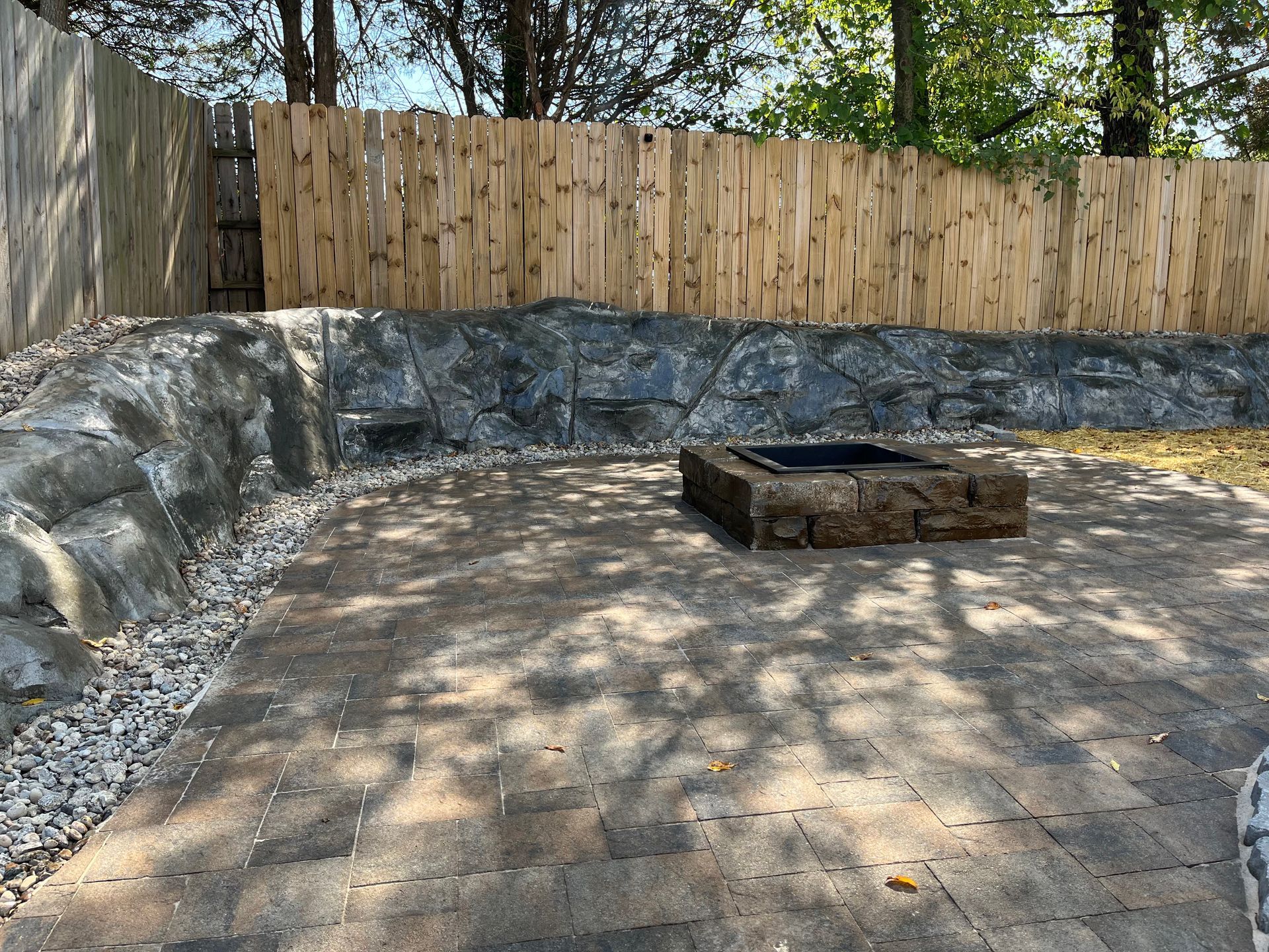 Stone-lined backyard fire pit area with a wooden fence and dappled sunlight on the ground