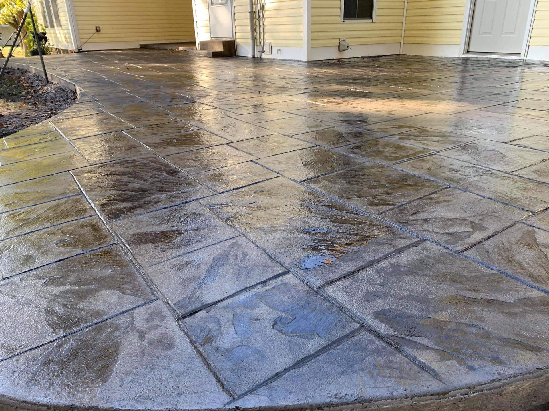 Wet stamped concrete patio outside a yellow house with white doors and a hose in the corner