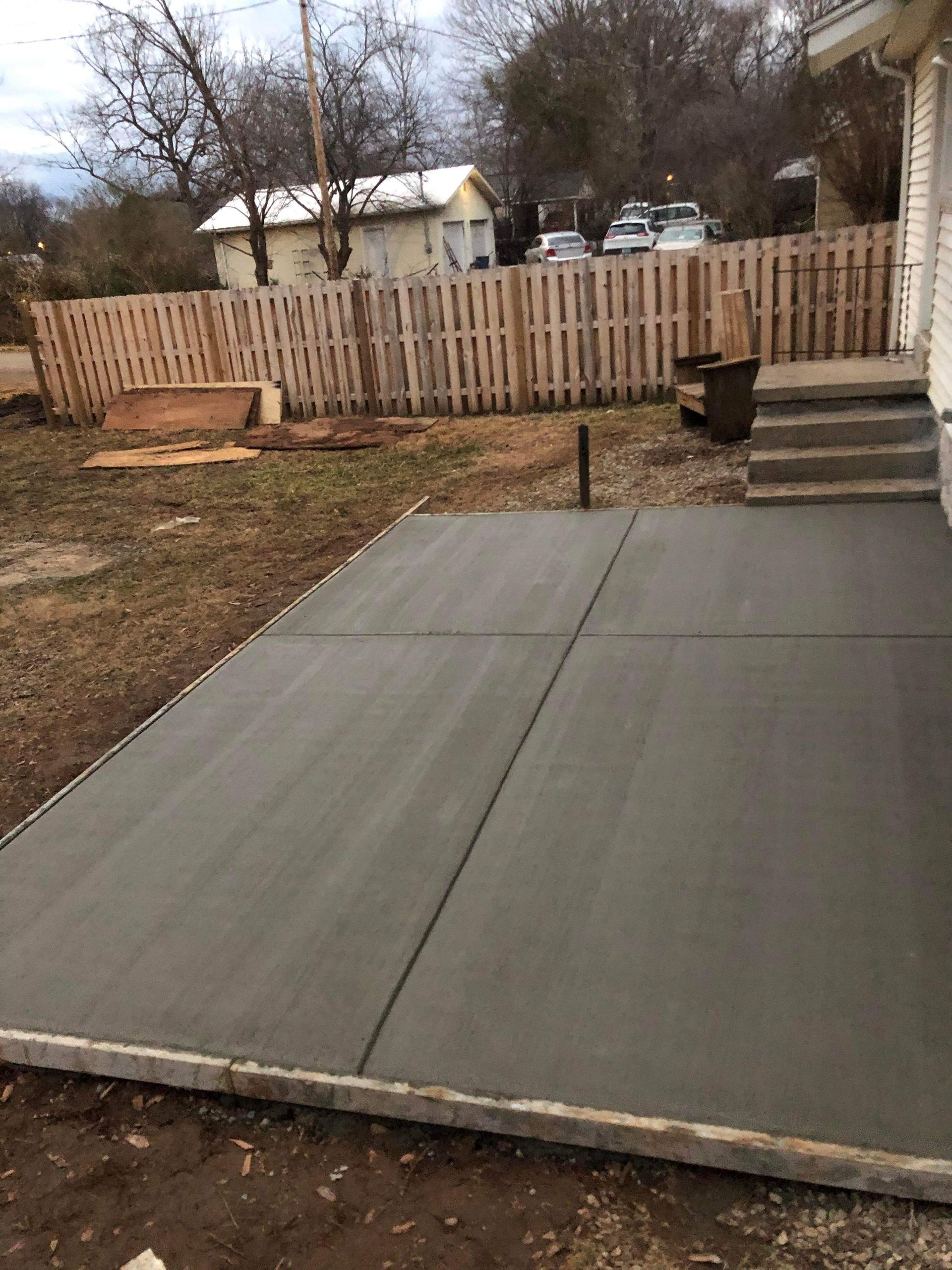 Freshly poured concrete patio slab in a backyard with a wooden fence and steps leading up to the yard