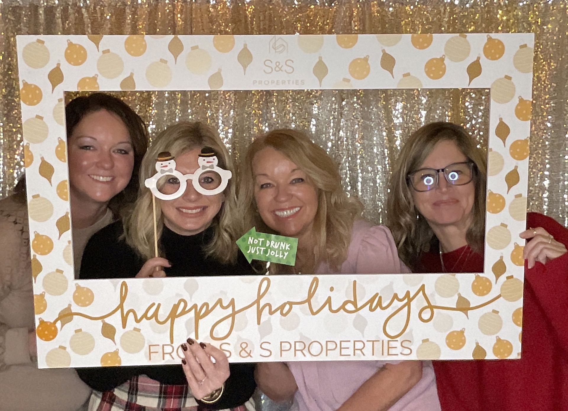 Four women pose smiling in a holiday photo frame at an event booth