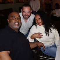 Three people smiling together at a dimly lit indoor gathering, seated close and posing for a photo.