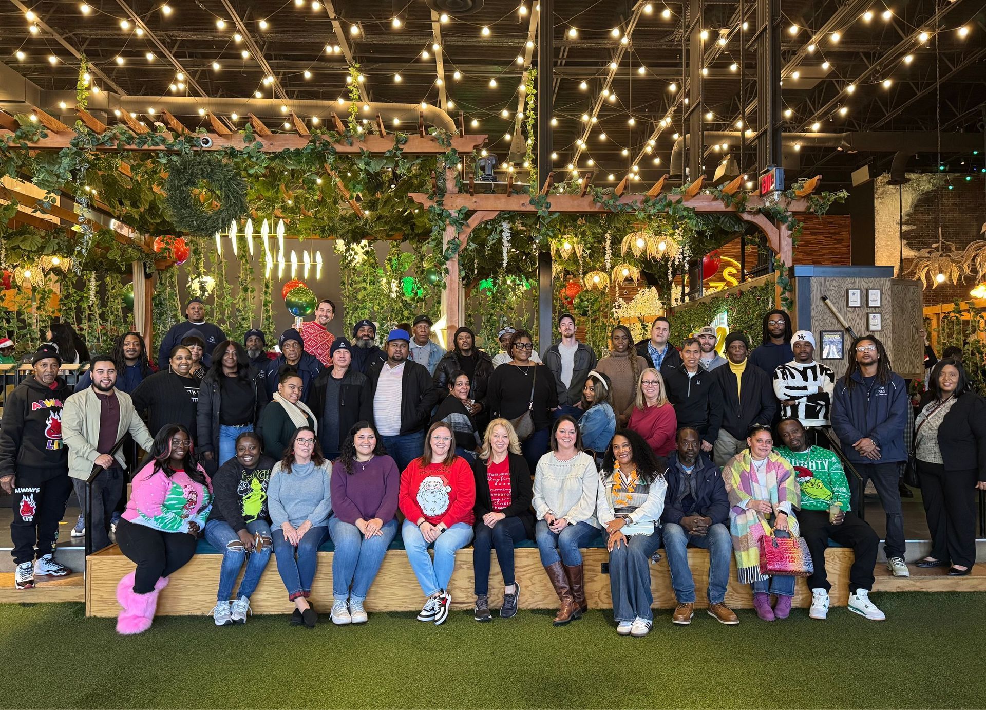 Large group posing indoors under string lights and greenery, seated and standing in front of a festive backdrop.