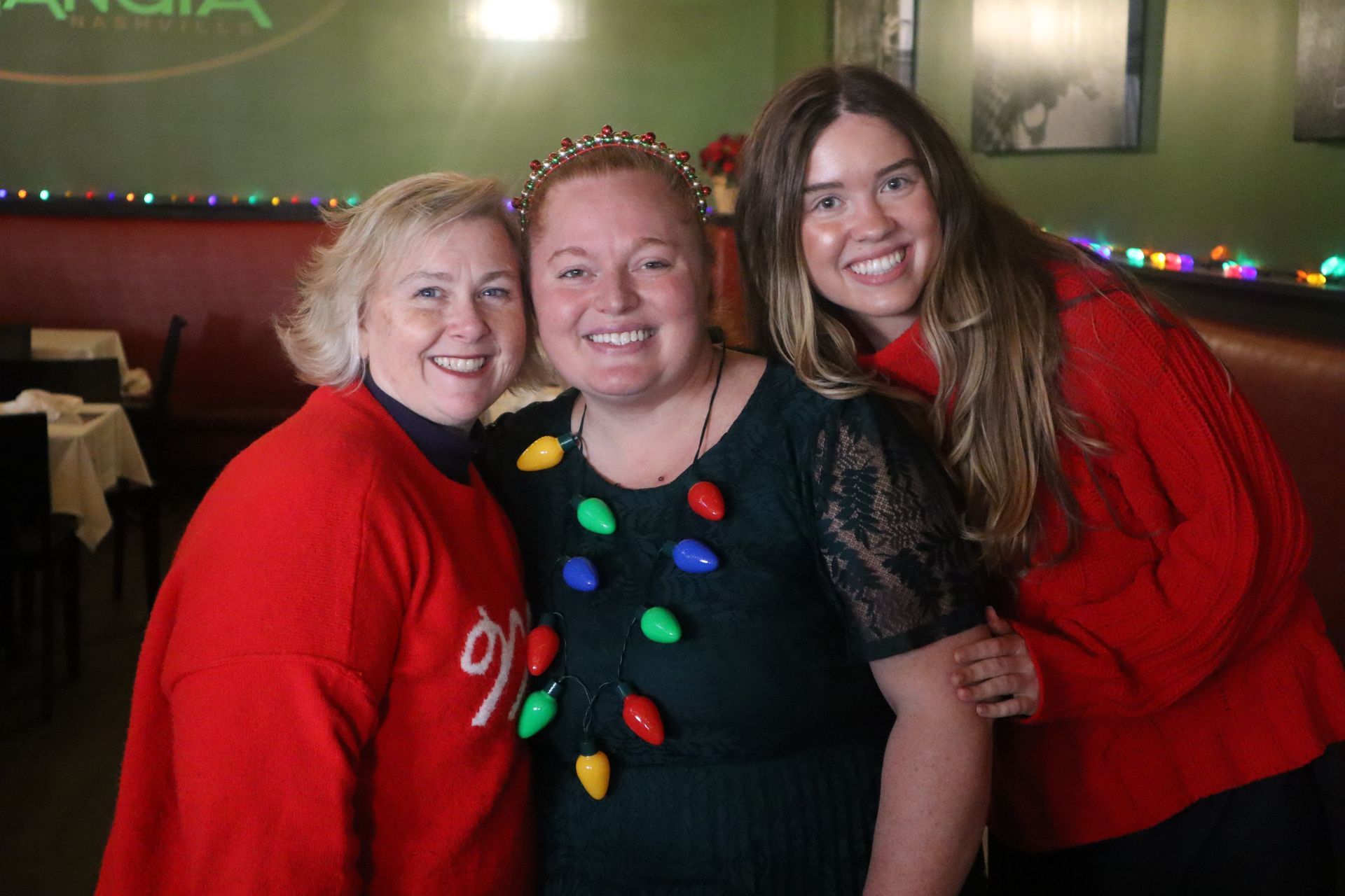 Three people smiling in red and black tops at a restaurant, with colorful string lights in the background.