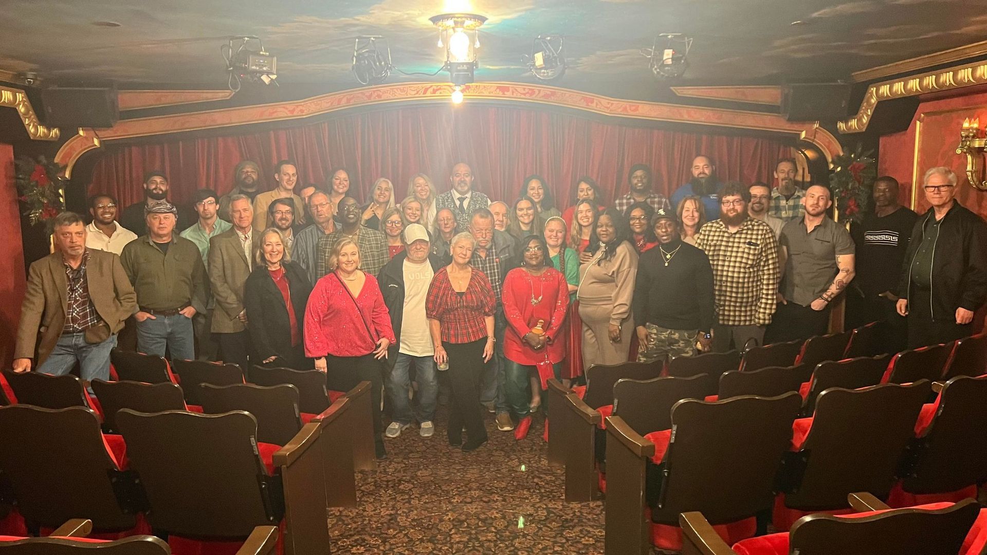 Group of people posing in a small theater, with red seats, curtain backdrop, and warm lighting