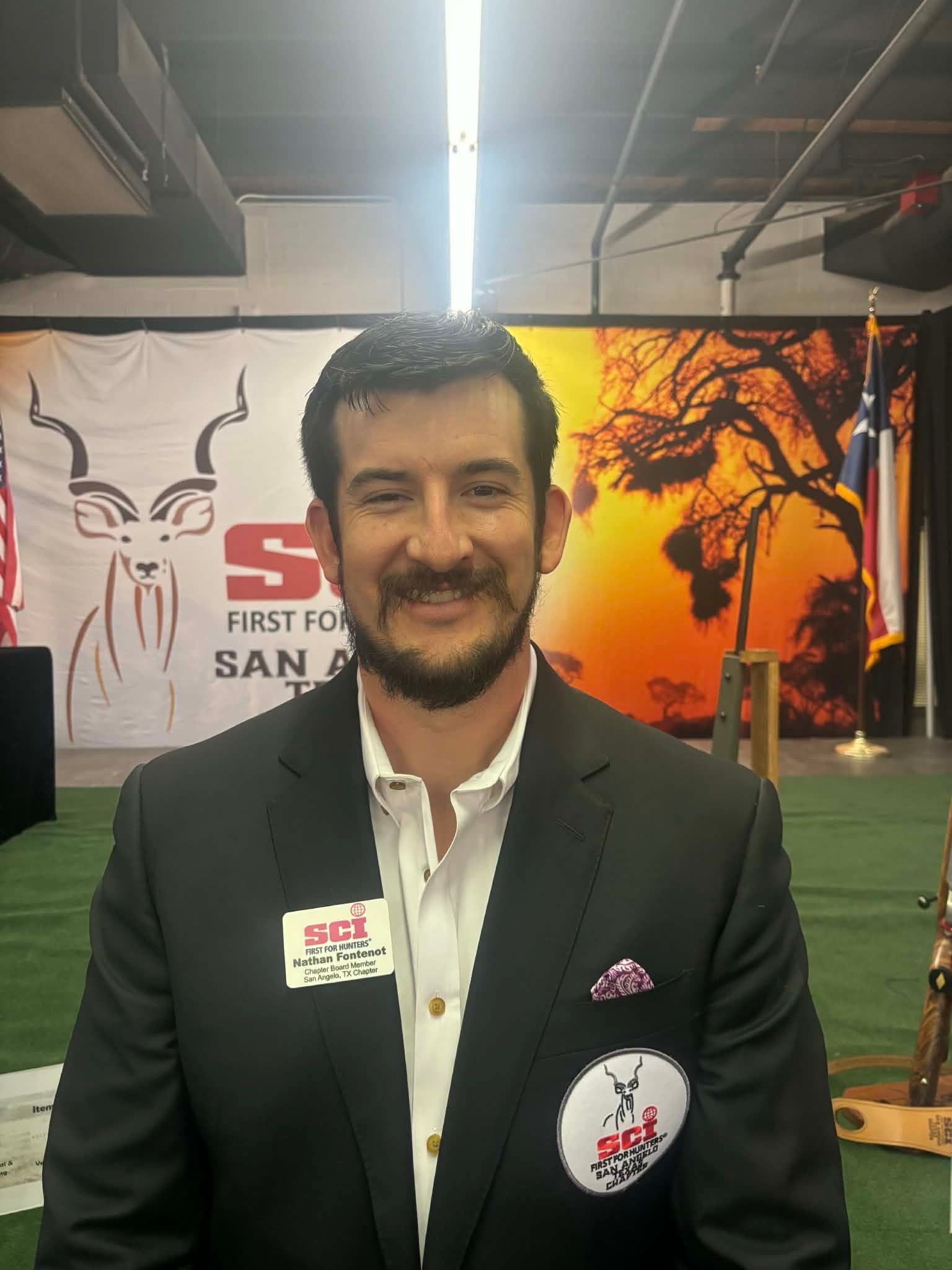 Man in dark suit standing at an indoor expo booth with a banner and bright overhead lights