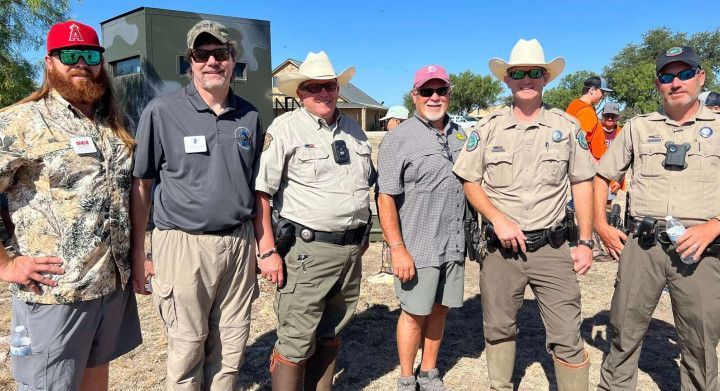 Six people in outdoor hats and uniforms posing together in bright sunlight before a rocky landscape