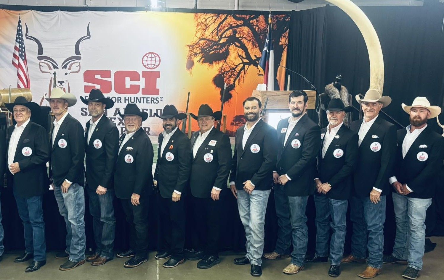 Group of people in matching blazers and cowboy hats posing onstage in front of a “SCI” banner.