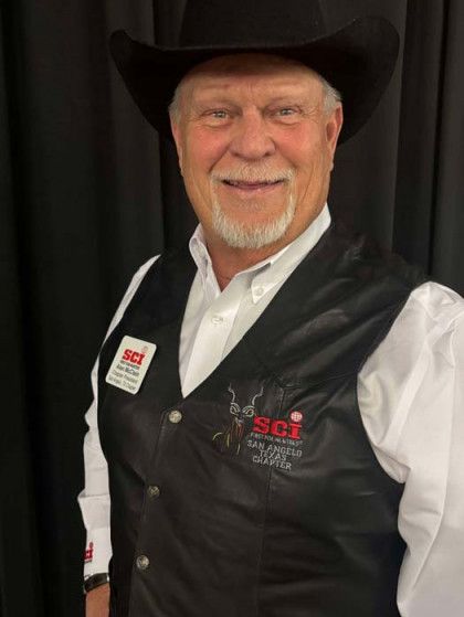Smiling man in a black cowboy hat and vest over a white shirt, standing against a dark background.