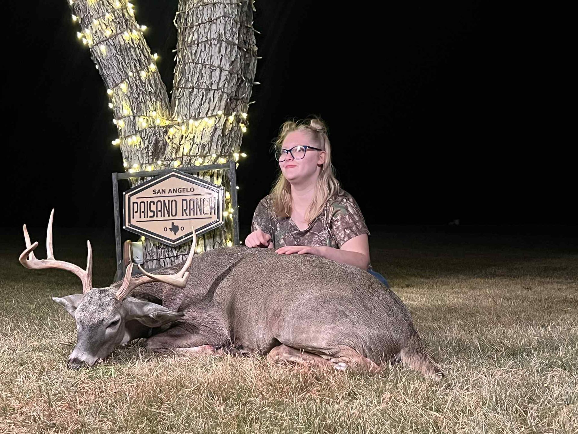 Hunter kneels beside a large deer at night near a lit sign and tree in a grassy field