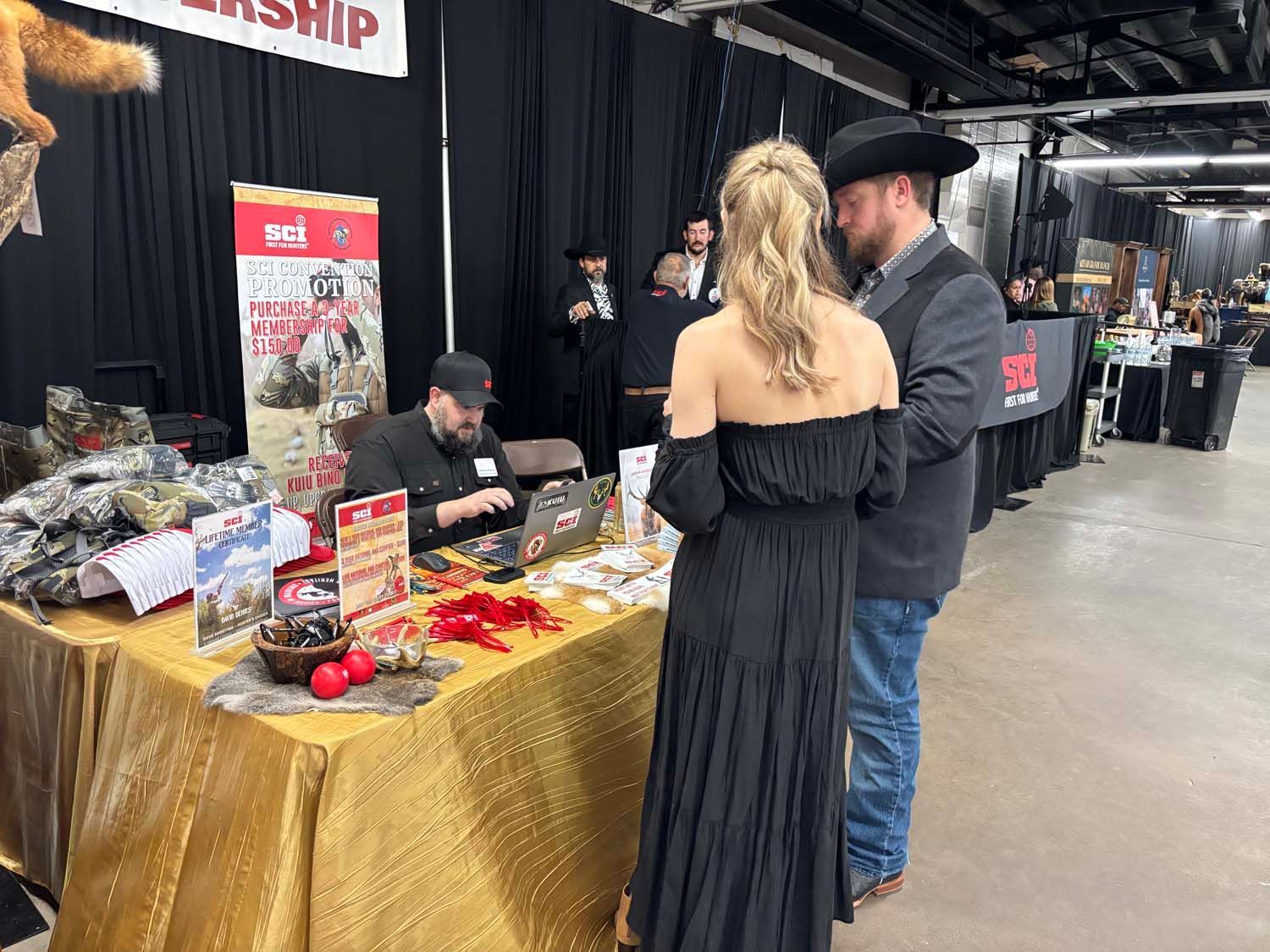 Market booth with gold tablecloth, red signs, and two people browsing at an indoor event