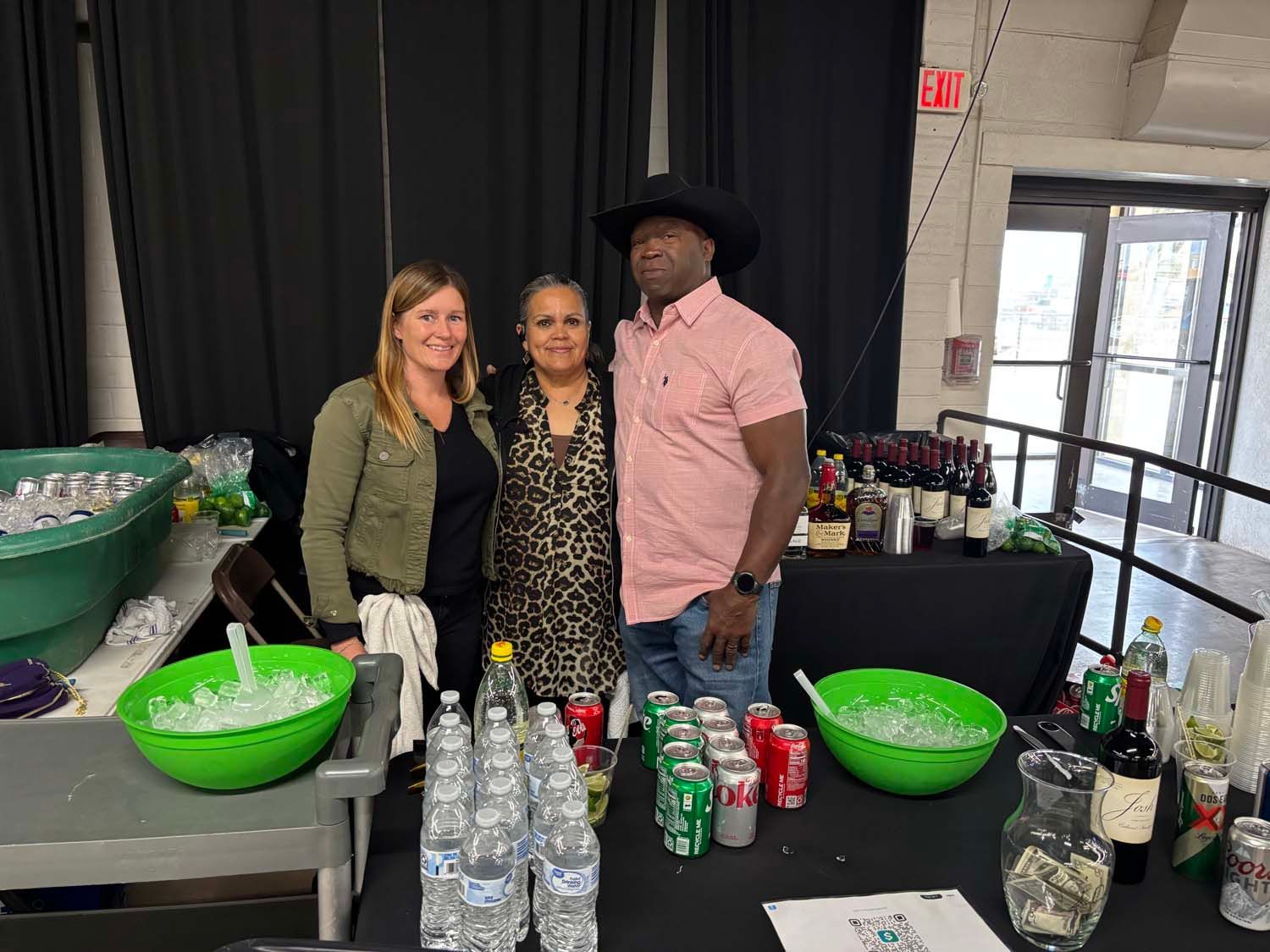 Three people standing behind a table of bottled drinks and green punch bowls at an indoor event