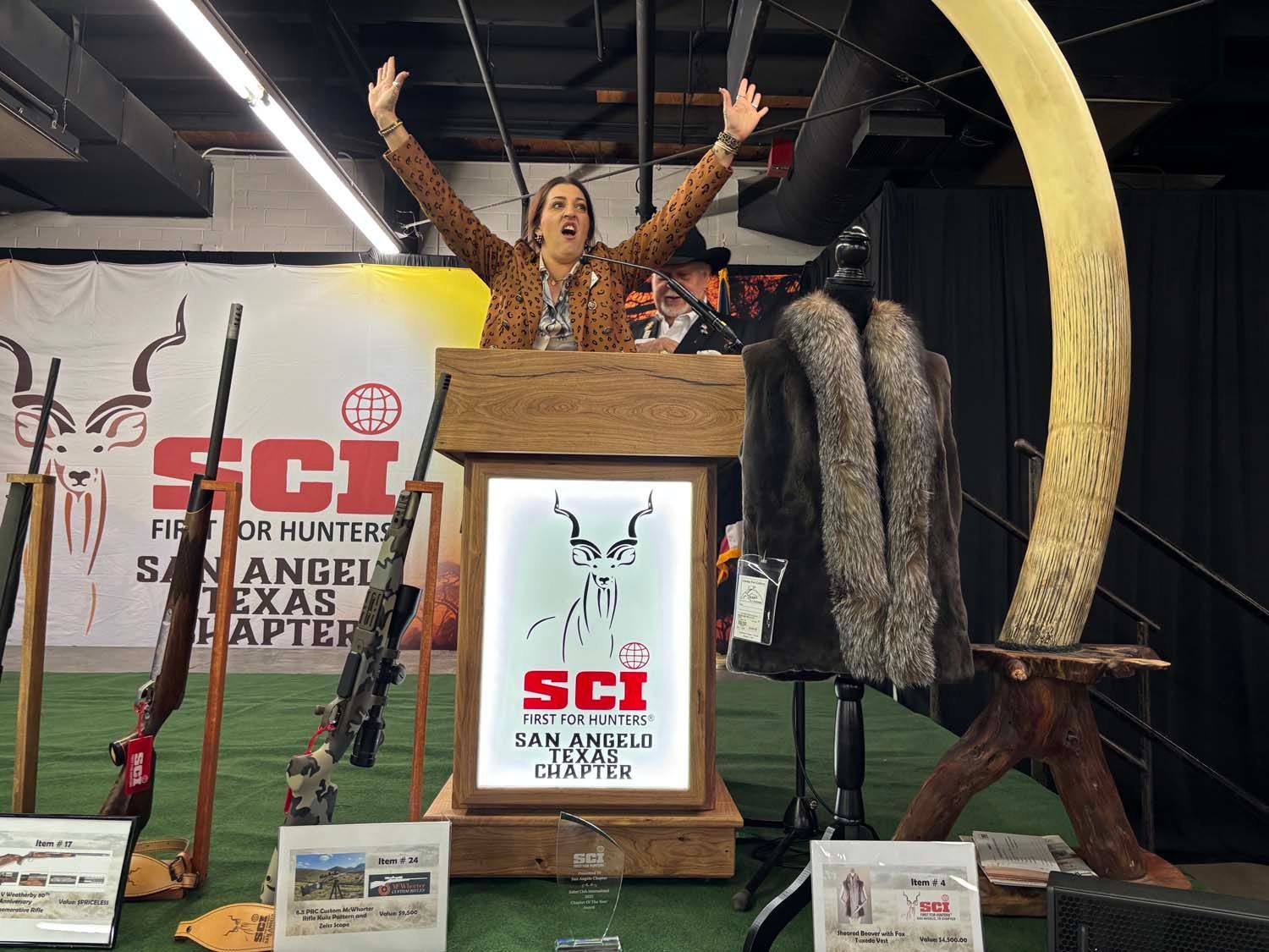 Woman posing on an SCI expo display with giant tusks and antlers, arms raised behind a podium.