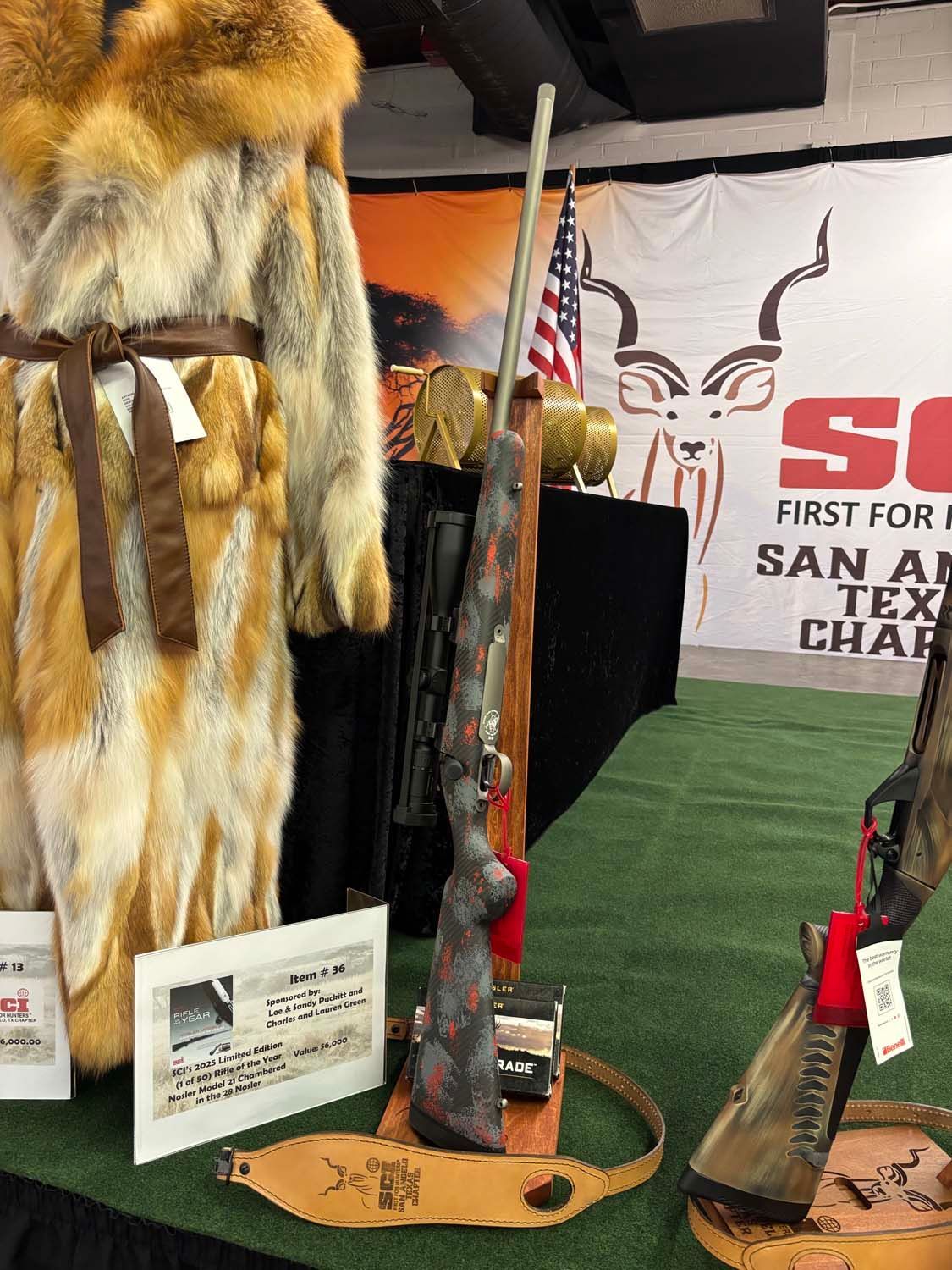 Display booth with fur coats, rifles, and hunting gear at a trade show in front of a San Antonio, Texas banner