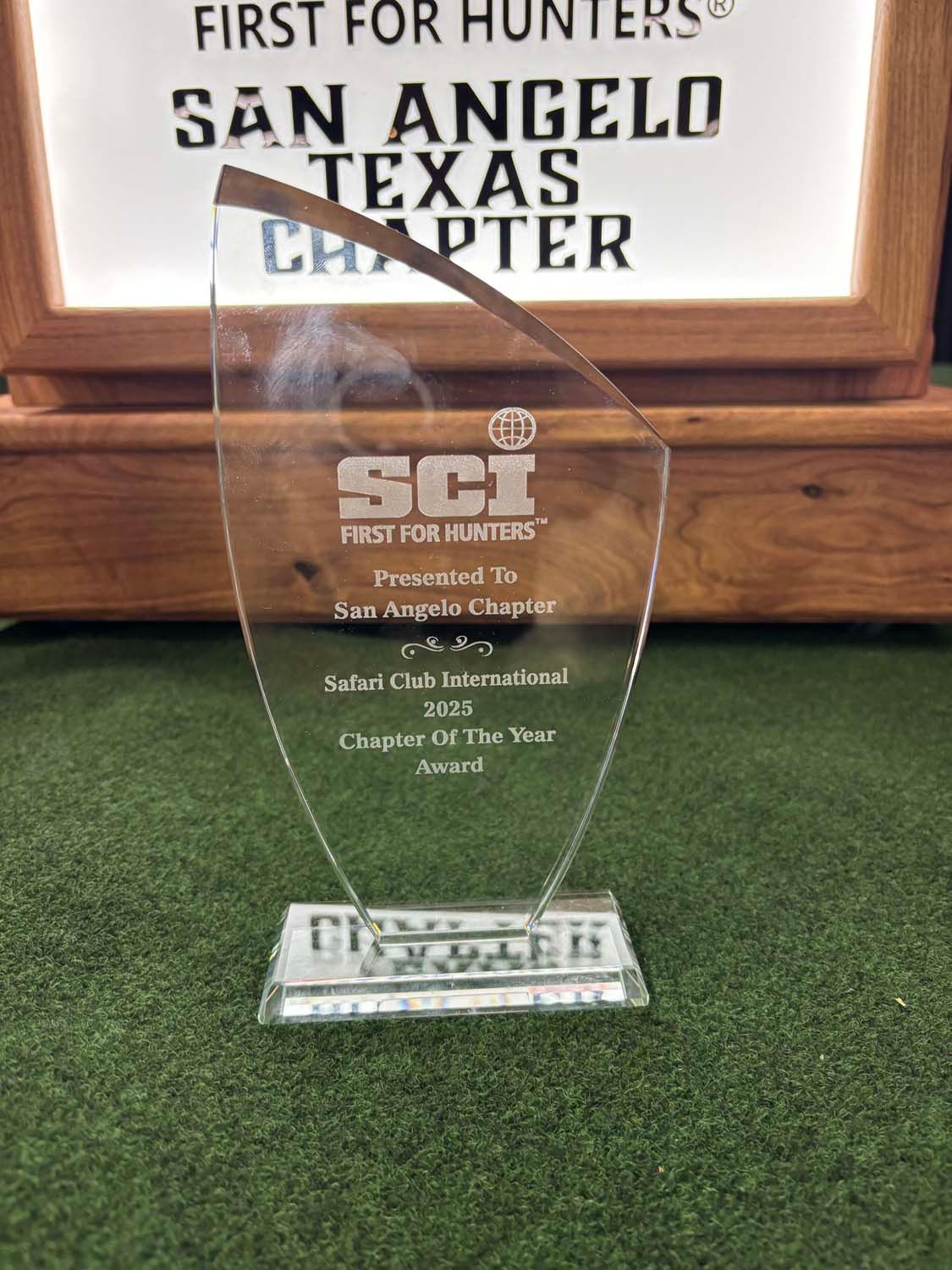Clear award plaque on green surface before a San Angelo, Texas chapter sign