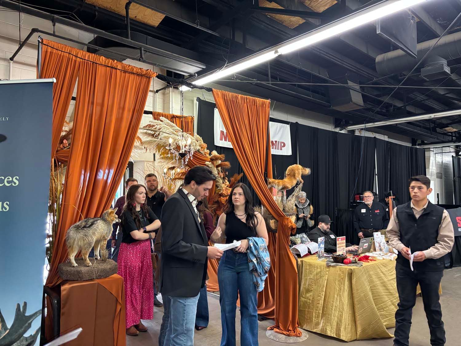 People gathered at a decorated event booth with orange drapes, a gold table, and a small animal statue display.