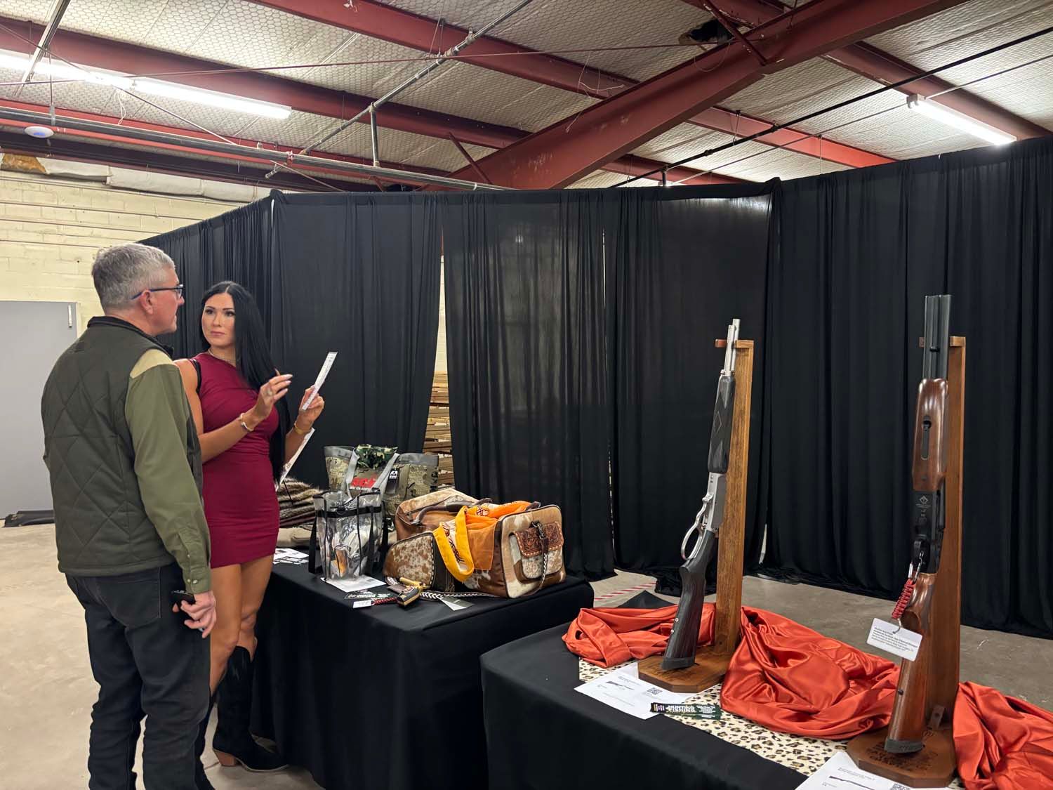 Two people talk beside an indoor display of long guns and orange cloth under black curtains.