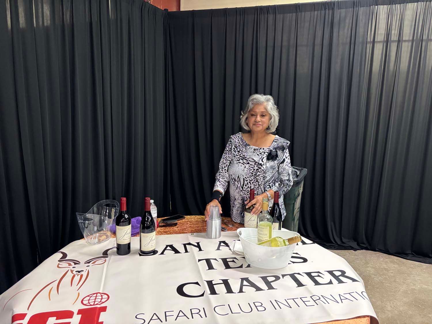 Woman behind a table with wine bottles and flowers in front of black curtains.