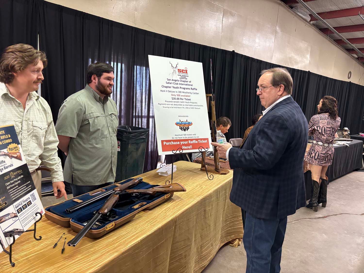 People at a booth displaying crossbows, with a man in a suit examining a sign at an indoor expo.