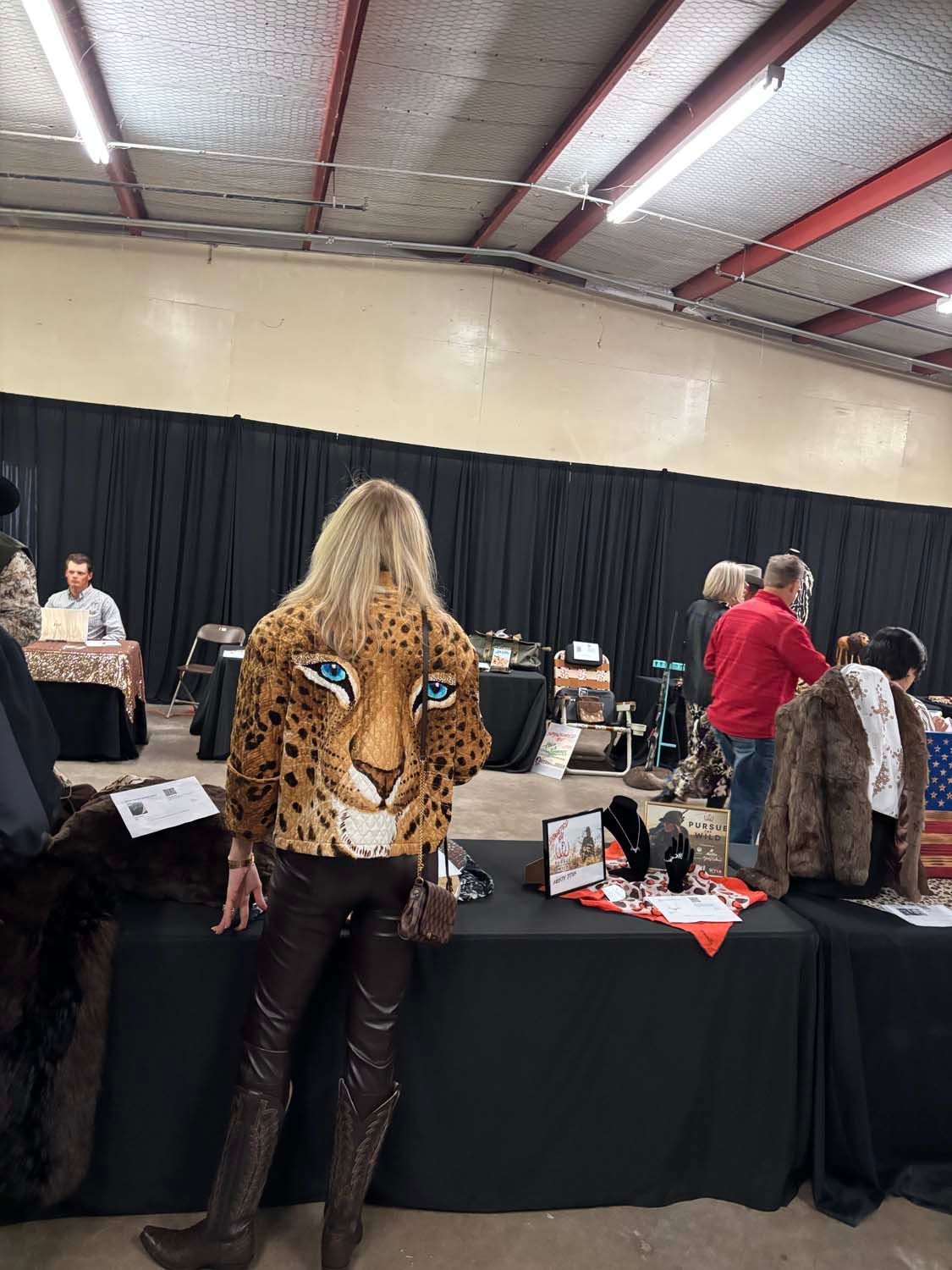 Vendor hall with black-draped tables and people browsing, centered on a leopard-print jacket on display.