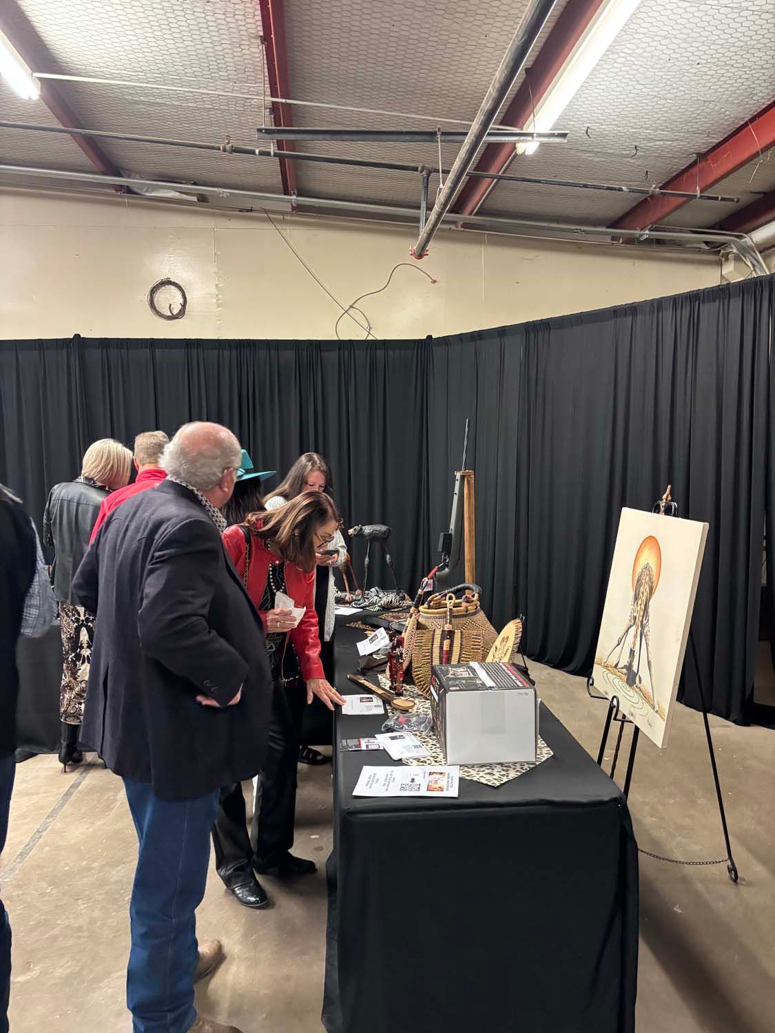 People browsing art displays at a table in a gallery booth with black curtains and a framed painting easel