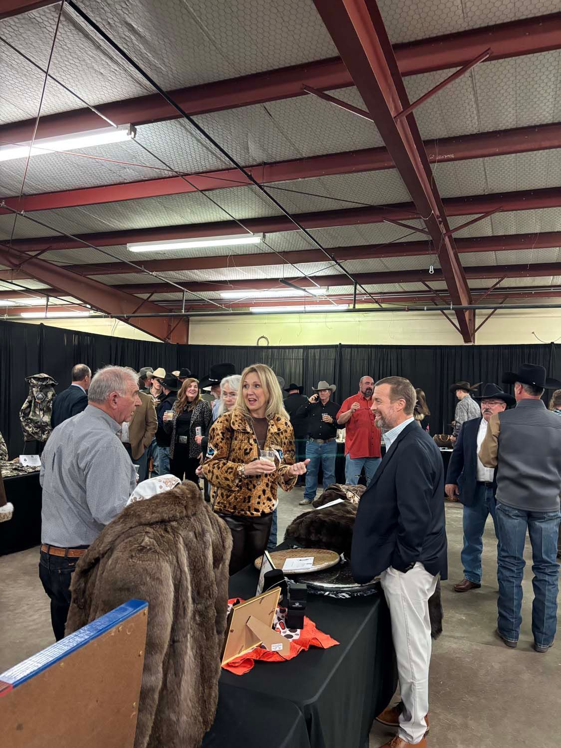 People chatting at an indoor event beneath a metal roof, with tables and black curtains in the background.