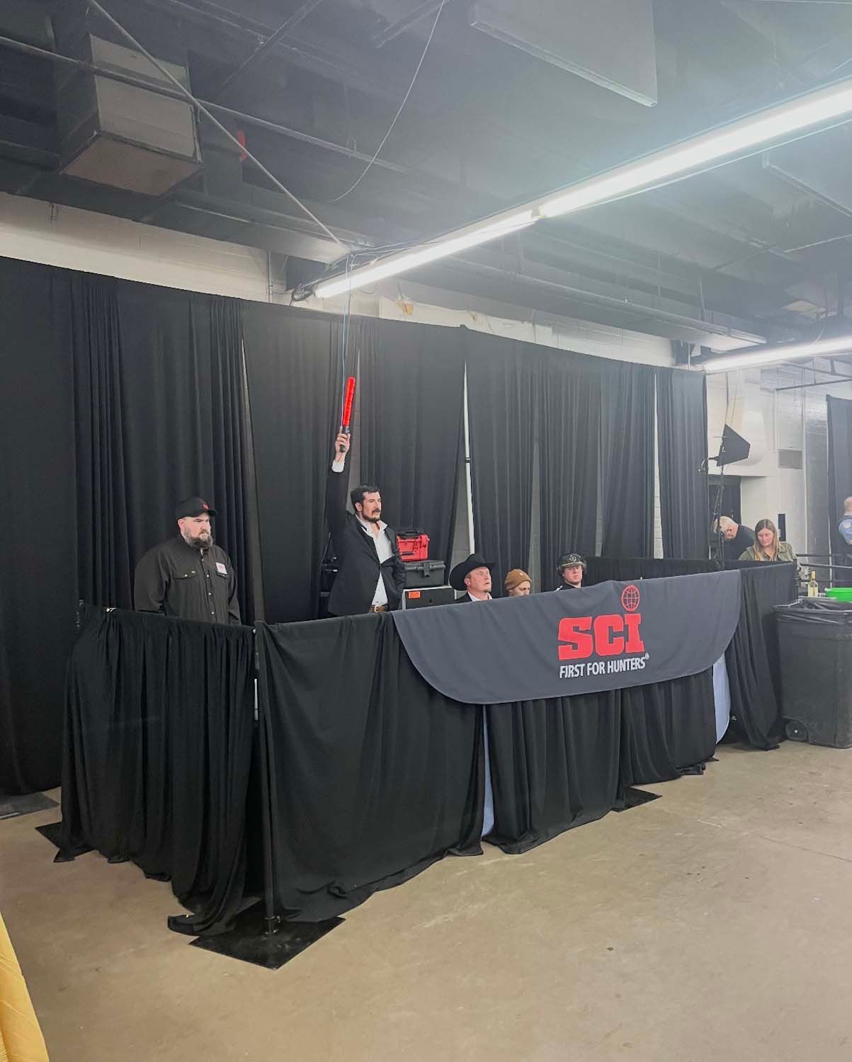 Three people seated behind a black SCI table at an indoor event stage with black drapes