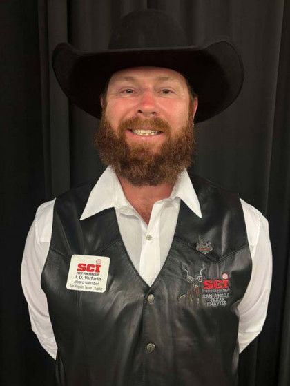 Smiling person in a black cowboy hat, white shirt, and black vest standing against a dark curtain.