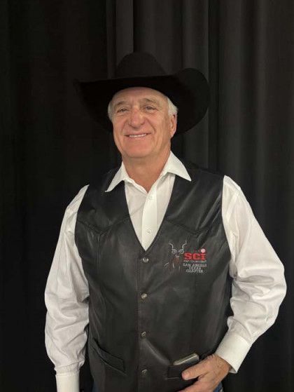 Smiling man in black cowboy hat and vest standing before a dark curtain