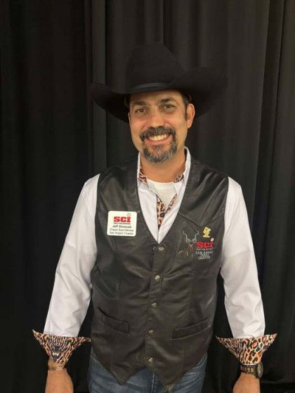 Man wearing black cowboy hat, white shirt, and black vest, smiling against a dark curtain backdrop