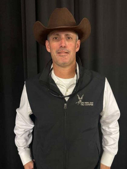 Man in a brown cowboy hat and black vest standing against a dark curtain backdrop.