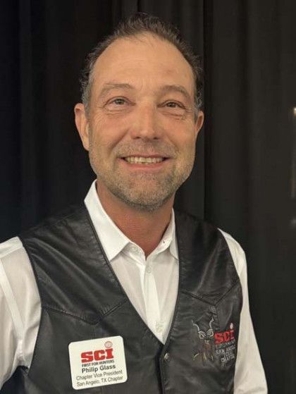 Smiling person in a white shirt and black vest with SCI logo, standing against a dark background