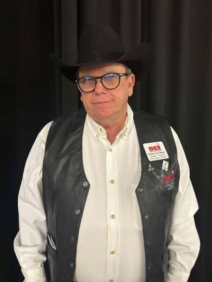 Man in black hat, glasses, white shirt, and black vest standing against a dark backdrop