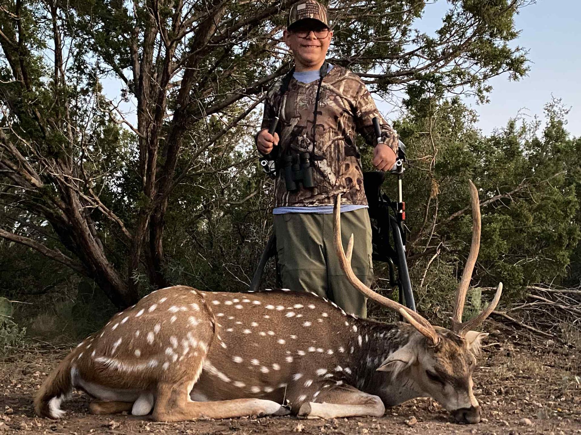 Hunter standing behind a spotted deer in a wooded area, holding a bow.