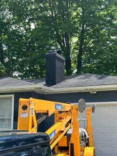 Yellow lift parked beside a house roof with a black chimney and tall trees behind it