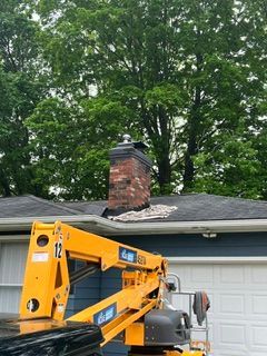 Yellow utility lift in a driveway beside a garage with a brick chimney and trees overhead