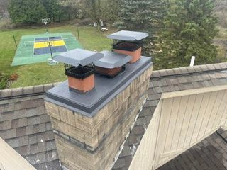 Rooftop chimneys with black caps on a brick house, overlooking a backyard with a trampoline and trees.